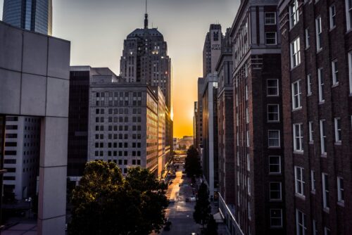 The image captures a cityscape at sunset, with golden light streaming between tall buildings. The architecture mixes modern and older styles, with sunlight reflecting off glass windows. Trees line the street below, and the sky transitions from deep blue to warm orange, creating a calm and serene atmosphere