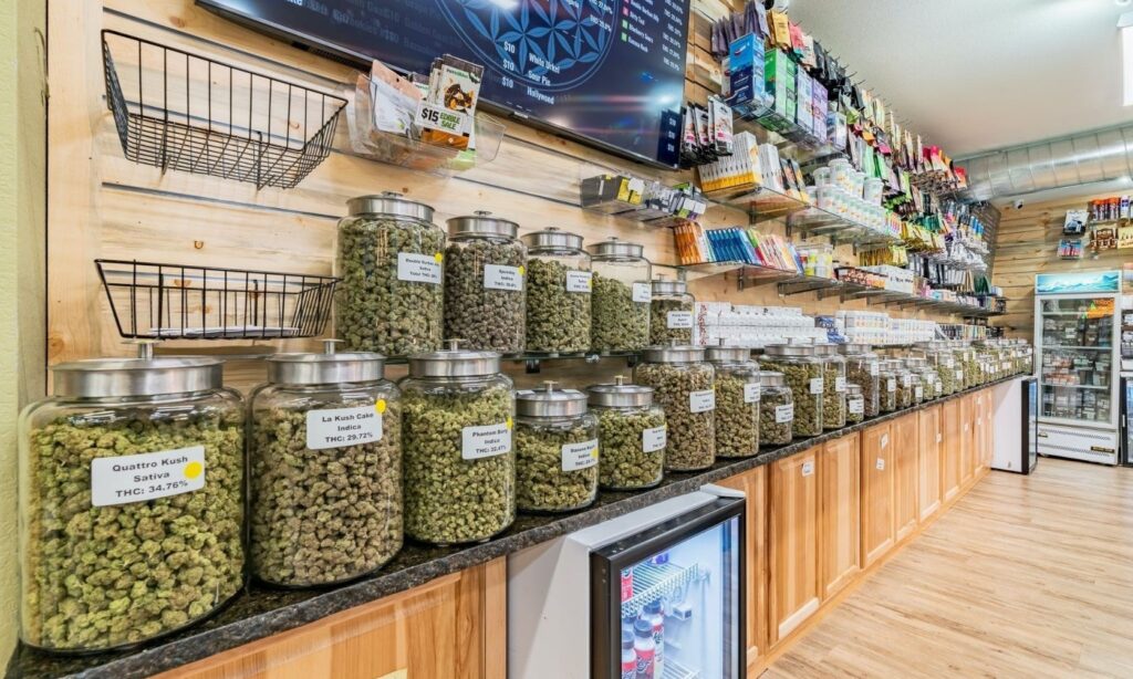 A well-stocked cannabis dispensary with jars of marijuana buds labeled with strain names and THC content, alongside various smoking accessories and edibles displayed on shelves.