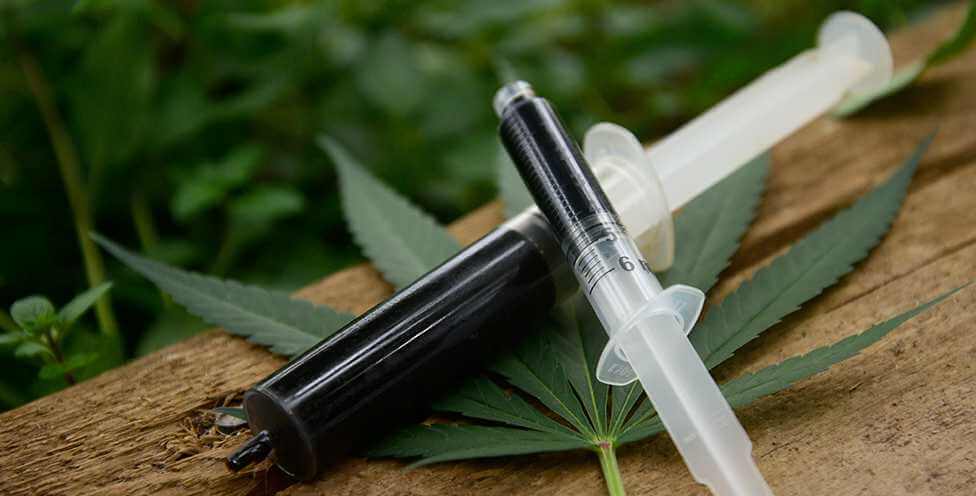 Two syringes, filled with Rick Simpson Oil (RSO), placed on a wooden surface with cannabis leaves in the background