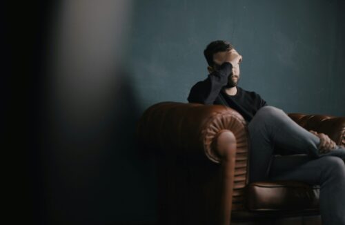 A man sits on a leather couch, covering his face with one hand, suggesting emotional distress or anxiety.