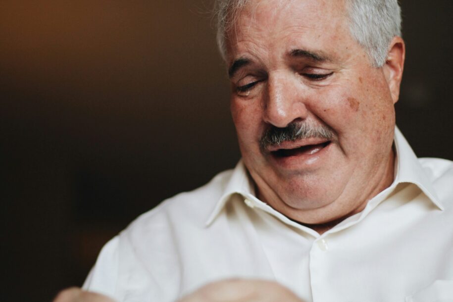 An elderly man with gray hair and a mustache, wearing a white shirt, appears emotional and teary-eyed, possibly reflecting sadness or distress.